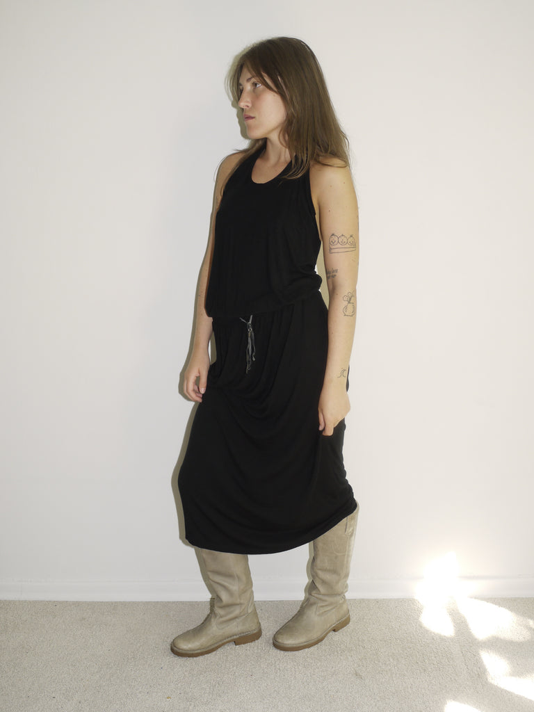 Model wearing a black sleeveless Diesel maxi dress with a relaxed fit and drawstring waist detail. Styled with chunky beige combat-style boots for a utilitarian edge. Shot against a plain white wall with natural light.