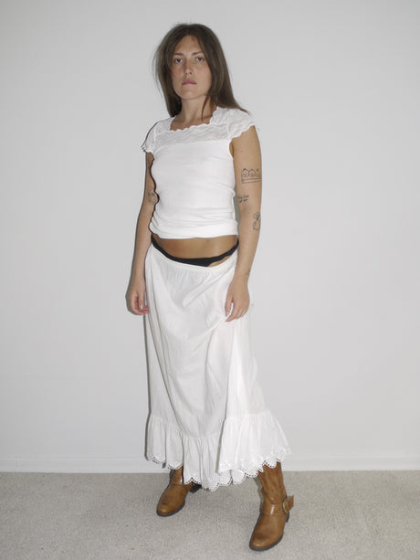 Model wearing a fitted white cap-sleeve top with lace detailing at the neckline, paired with a low-rise white skirt featuring ruffled hem and lace trim. Styled with brown leather Western boots for a romantic-meets-rugged aesthetic. Shot against a plain white wall.