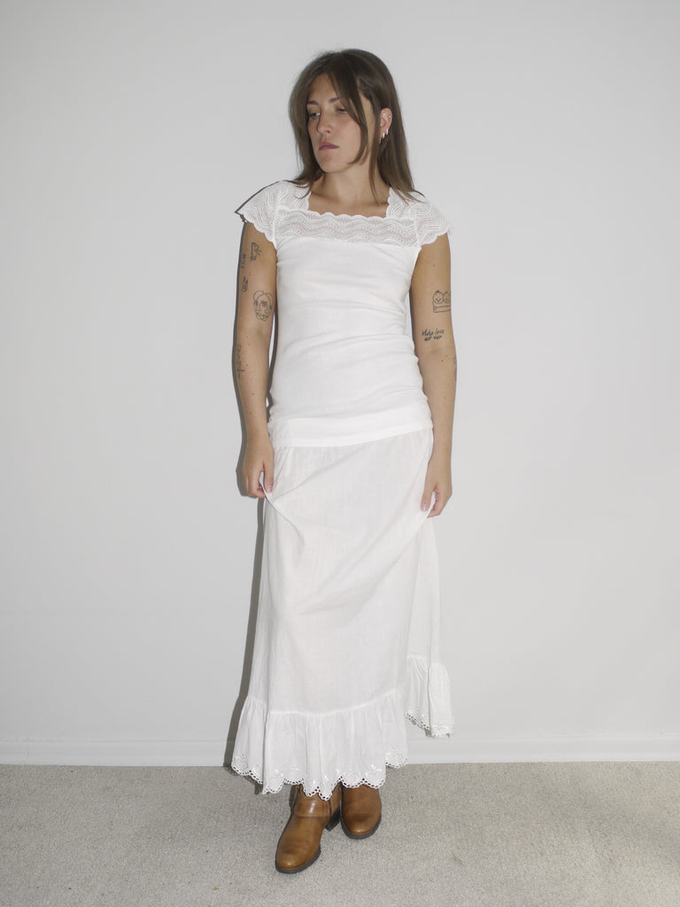 Model wearing a fitted white cap-sleeve top with lace detailing at the neckline, paired with a low-rise white skirt featuring ruffled hem and lace trim. Styled with brown leather Western boots for a romantic-meets-rugged aesthetic. Shot against a plain white wall.