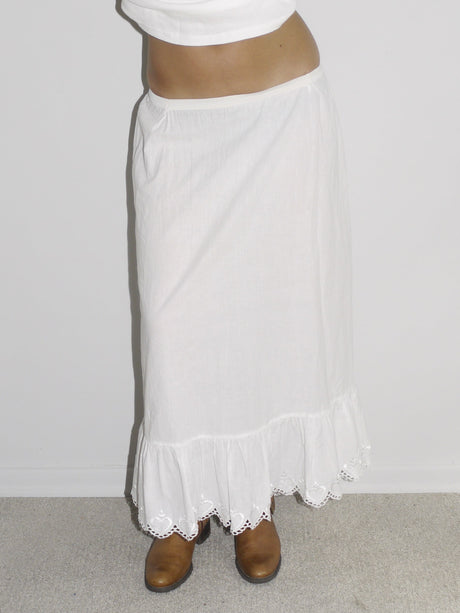 Model wearing a fitted white cap-sleeve top with lace detailing at the neckline, paired with a low-rise white skirt featuring ruffled hem and lace trim. Styled with brown leather Western boots for a romantic-meets-rugged aesthetic. Shot against a plain white wall.