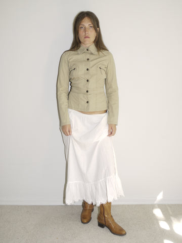 Model wearing a fitted khaki button-up jacket with chest pockets, paired with a white low-rise skirt featuring ruffle hem and lace trim. Styled with tall tan Western boots for a vintage prairie-inspired look. Shot against a plain white wall with soft natural light