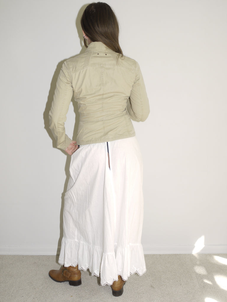 Model wearing a fitted khaki button-up jacket with chest pockets, paired with a white low-rise skirt featuring ruffle hem and lace trim. Styled with tall tan Western boots for a vintage prairie-inspired look. Shot against a plain white wall with soft natural light