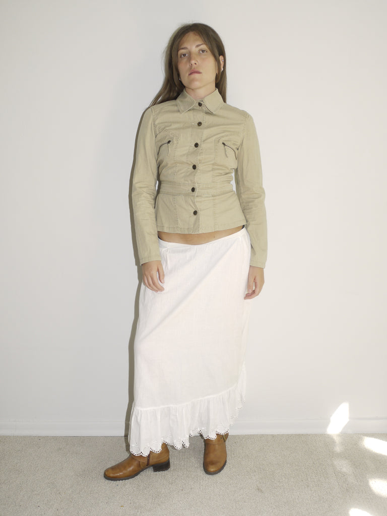 Model wearing a fitted khaki button-up jacket with chest pockets, paired with a white low-rise skirt featuring ruffle hem and lace trim. Styled with tall tan Western boots for a vintage prairie-inspired look. Shot against a plain white wall with soft natural light