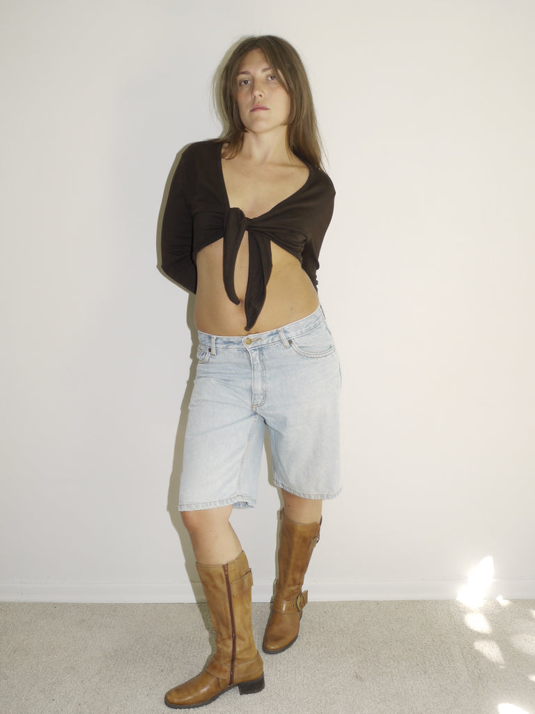  view of a model wearing a black long-sleeve crop top and light-wash vintage denim Bermuda shorts with a visible leather patch label. Styled with tall tan Western boots featuring gold-tone buckles. Shot against a plain white wall with soft natural lighting.