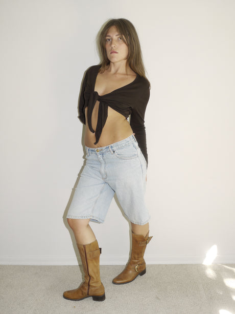  view of a model wearing a black long-sleeve crop top and light-wash vintage denim Bermuda shorts with a visible leather patch label. Styled with tall tan Western boots featuring gold-tone buckles. Shot against a plain white wall with soft natural lighting.