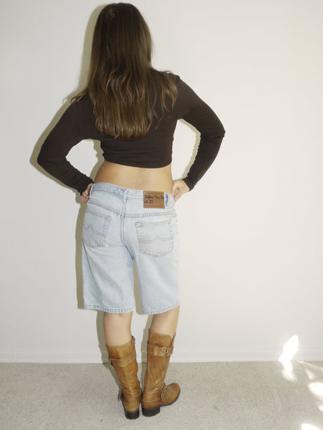 Back view of a model wearing a black long-sleeve crop top and light-wash vintage denim Bermuda shorts with a visible leather patch label. Styled with tall tan Western boots featuring gold-tone buckles. Shot against a plain white wall with soft natural lighting.