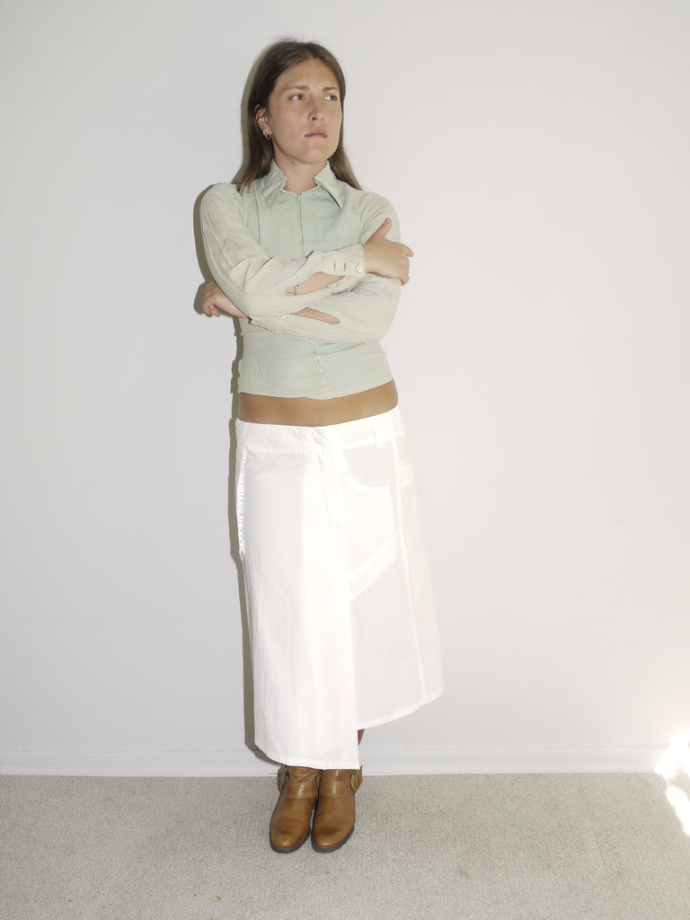 Model wearing a fitted pale green button-up top with long sleeves and a slightly cropped cut, paired with a low-rise white midi skirt with flowy silhouette and tie detail at the waist. Styled with tall tan Western boots. Shot against a white wall with soft natural lighting