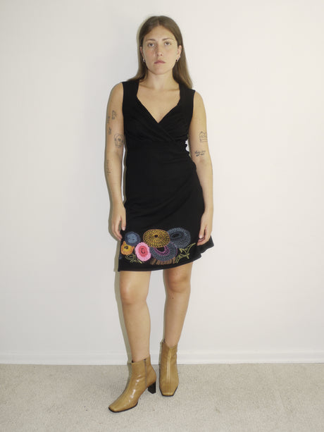 Model wearing a sleeveless black Desigual dress with a deep V-neckline and colorful floral embroidery along the hem. Styled with tan ankle boots for a bold contrast between playful detailing and minimal silhouette. Shot against a plain white background