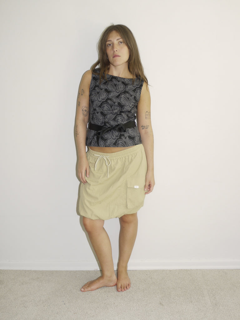 Model wearing a sleeveless fitted top with black floral jacquard pattern, paired with a low-rise beige mini skirt featuring drawstring details and a sporty silhouette. Barefoot styling gives it a casual, lived-in feel. Shot against a plain white wall.