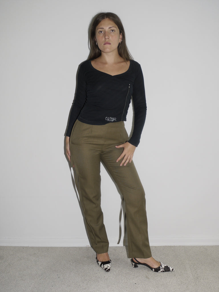 Model wearing a long-sleeve black top with asymmetrical seams, paired with iconic olive green Maison Margiela pants featuring deconstructed strap detailing hanging from the waistband. Styled with black-and-white printed heels for a conceptual, avant-garde look. Shot against a plain white background.