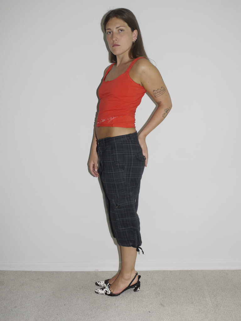 Model wearing a fitted red tank top with subtle graphic detailing, paired with low-rise dark capri pants featuring a checkered pattern and drawstring hems. Styled with black-and-white printed slingback heels, creating a sharp Y2K-inspired contrast. Shot against a plain white wall.