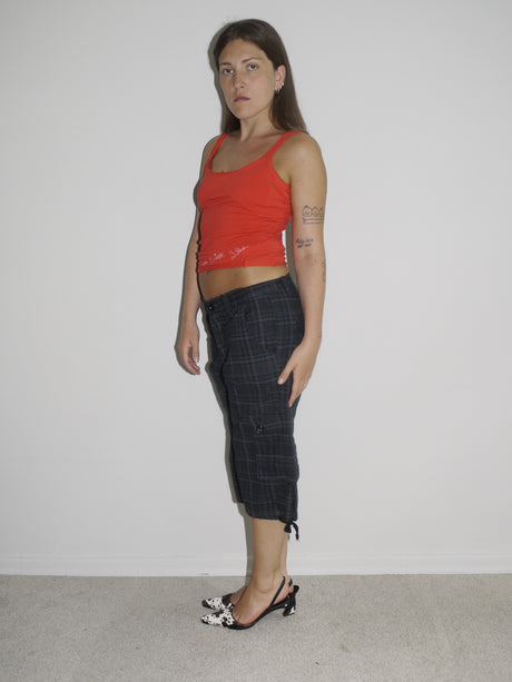 Model wearing a fitted red tank top with subtle graphic detailing, paired with low-rise dark capri pants featuring a checkered pattern and drawstring hems. Styled with black-and-white printed slingback heels, creating a sharp Y2K-inspired contrast. Shot against a plain white wall.