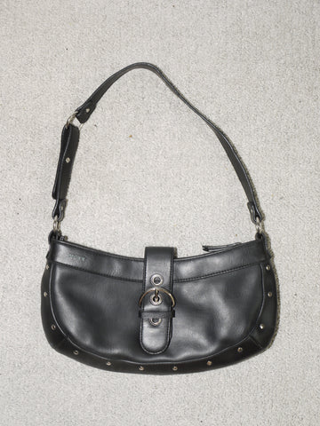 Vintage black leather shoulder bag with silver-tone hardware, buckle front detail, and stud accents along the edges. Features a short strap and a zip closure, evoking early 2000s It-bag energy. Laid flat on a neutral carpet background.