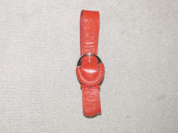 Vintage red leather belt with a glossy finish, featuring a round metal buckle wrapped in matching red leather detailing. Laid flat on a neutral carpet background. Bold and retro-inspired.