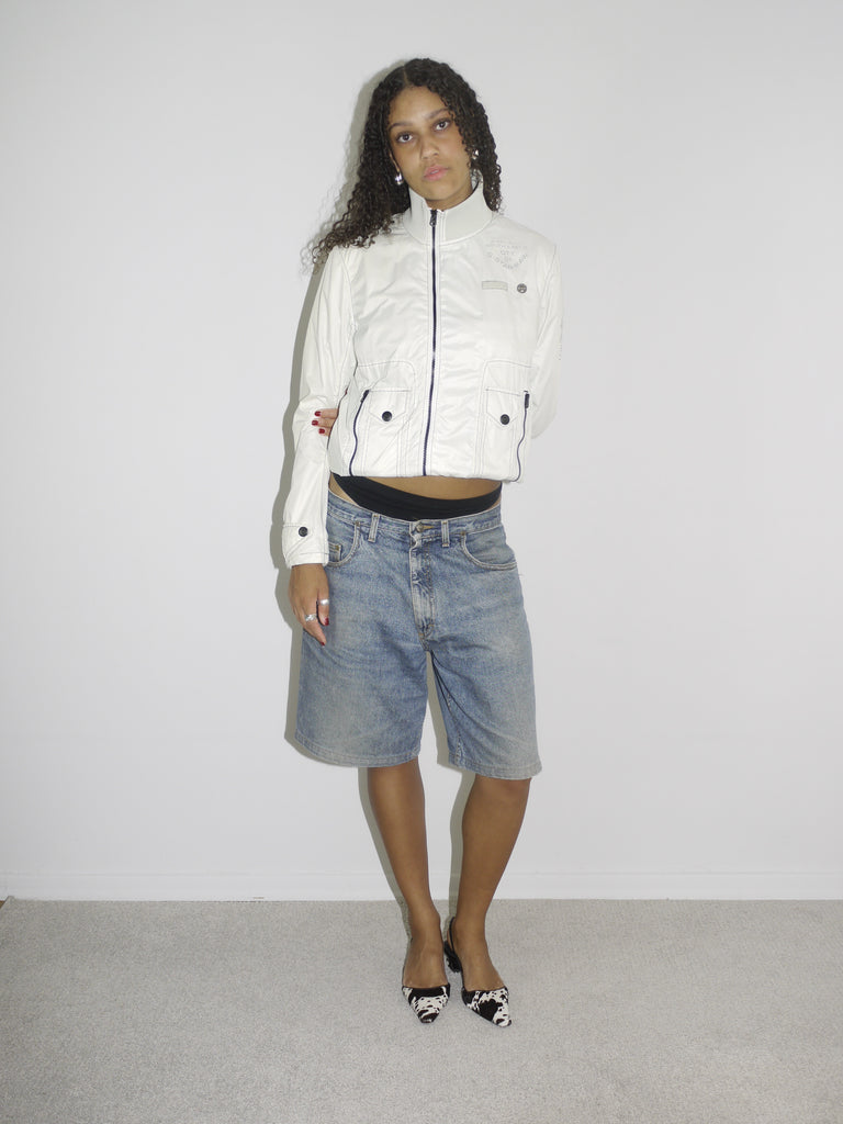 Model wearing a structured white zip-up jacket with black trim and button details, paired with loose-fitting vintage denim Bermuda shorts. Styled with pointed black-and-white printed slingback heels, blending utilitarian outerwear with early-2000s femininity. Shot against a plain white wall.