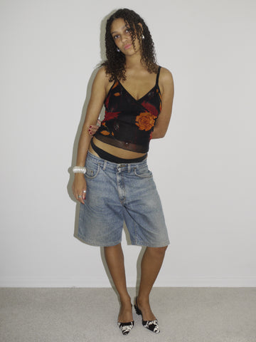Model wearing a sheer black mesh camisole with red and orange floral embroidery and side cut-outs, paired with low-rise vintage denim Bermuda shorts. Styled with pointed black-and-white printed heels and silver jewelry for a bold Y2K-inspired going-out look. Shot against a plain white wall.