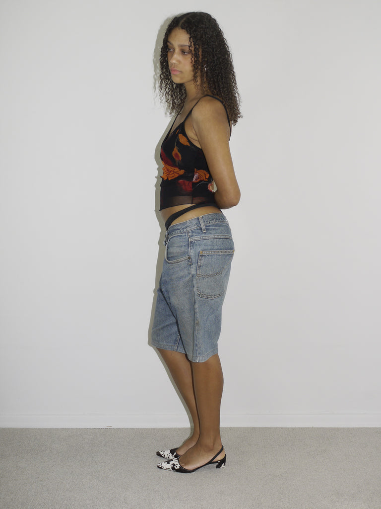 Model wearing a sheer black mesh camisole with red and orange floral embroidery and side cut-outs, paired with low-rise vintage denim Bermuda shorts. Styled with pointed black-and-white printed heels and silver jewelry for a bold Y2K-inspired going-out look. Shot against a plain white wall.