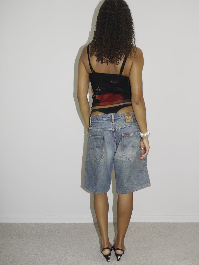 Model wearing a sheer black mesh camisole with red and orange floral embroidery and side cut-outs, paired with low-rise vintage denim Bermuda shorts. Styled with pointed black-and-white printed heels and silver jewelry for a bold Y2K-inspired going-out look. Shot against a plain white wall.