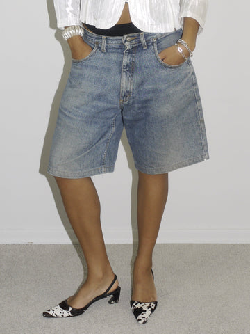 Model wearing a structured white zip-up jacket with black trim and button details, paired with loose-fitting vintage denim Bermuda shorts. Styled with pointed black-and-white printed slingback heels, blending utilitarian outerwear with early-2000s femininity. Shot against a plain white wall.
