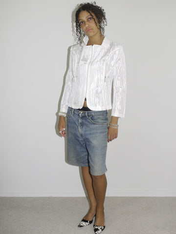 Model wearing a shiny textured white blazer with a subtle sheen, styled over low-rise vintage denim Bermuda shorts. Finished with black-and-white pointed heels and delicate jewelry, combining business-core elements with laid-back Y2K denim. Shot against a plain white background.