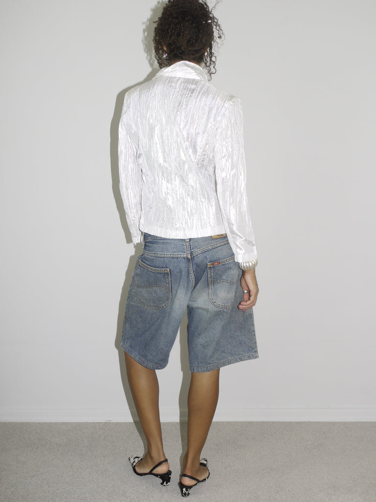 Model wearing a shiny textured white blazer with a subtle sheen, styled over low-rise vintage denim Bermuda shorts. Finished with black-and-white pointed heels and delicate jewelry, combining business-core elements with laid-back Y2K denim. Shot against a plain white background.