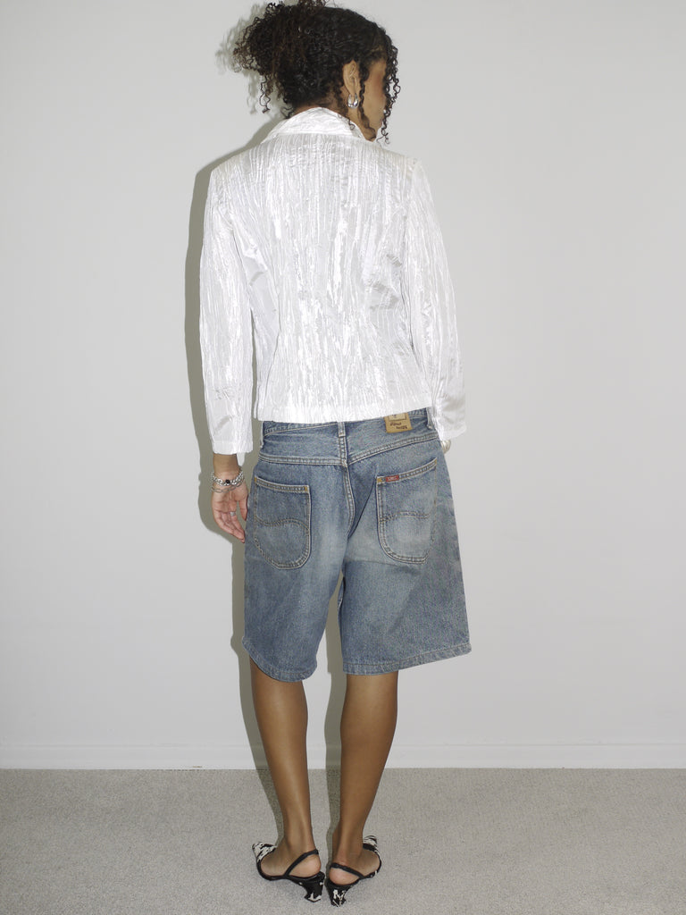 Model wearing a shiny textured white blazer with a subtle sheen, styled over low-rise vintage denim Bermuda shorts. Finished with black-and-white pointed heels and delicate jewelry, combining business-core elements with laid-back Y2K denim. Shot against a plain white background.