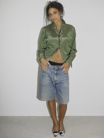 Model wearing a crinkled olive green button-up shirt, left slightly unbuttoned to reveal a black waistband, paired with loose-fitting vintage denim Bermuda shorts. Styled with pointed black-and-white printed heels for a contrast of grunge and glam. Shot against a plain white background.