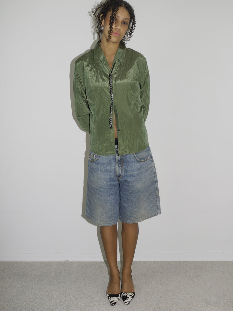 Model wearing a crinkled olive green button-up shirt, left slightly unbuttoned to reveal a black waistband, paired with loose-fitting vintage denim Bermuda shorts. Styled with pointed black-and-white printed heels for a contrast of grunge and glam. Shot against a plain white background.