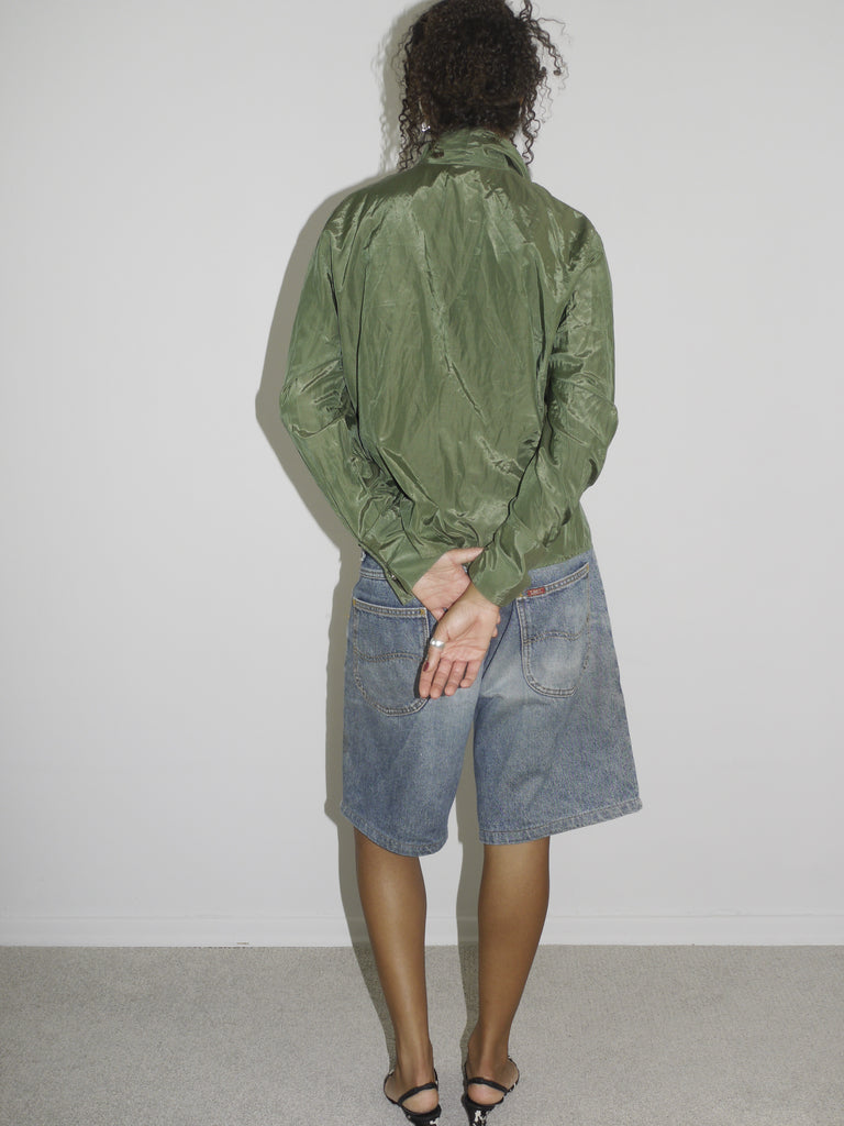 Model wearing a crinkled olive green button-up shirt, left slightly unbuttoned to reveal a black waistband, paired with loose-fitting vintage denim Bermuda shorts. Styled with pointed black-and-white printed heels for a contrast of grunge and glam. Shot against a plain white background.