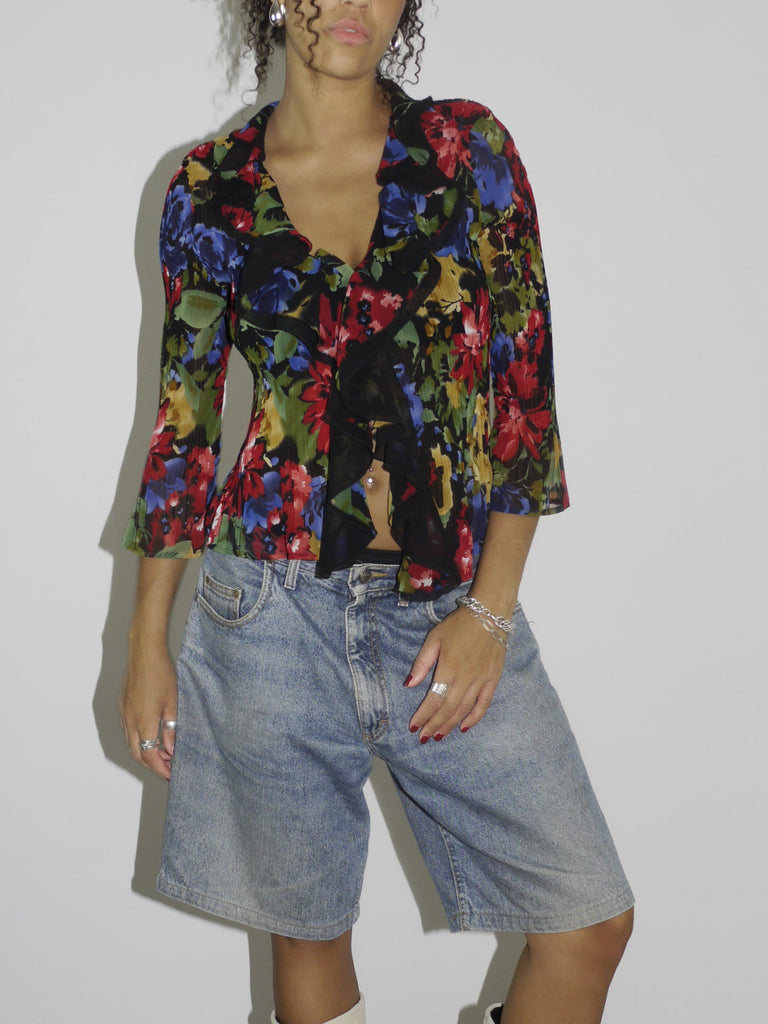 Model wearing a colorful sheer floral wrap blouse with a deep neckline, paired with loose-fitting vintage denim Bermuda shorts. Styled with cream-colored pointed cowboy boots and silver jewelry, blending romantic boho elements with early-2000s streetwear. Shot against a plain white wall