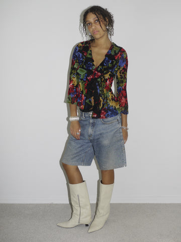 Model wearing a colorful sheer floral wrap blouse with a deep neckline, paired with loose-fitting vintage denim Bermuda shorts. Styled with cream-colored pointed cowboy boots and silver jewelry, blending romantic boho elements with early-2000s streetwear. Shot against a plain white wall
