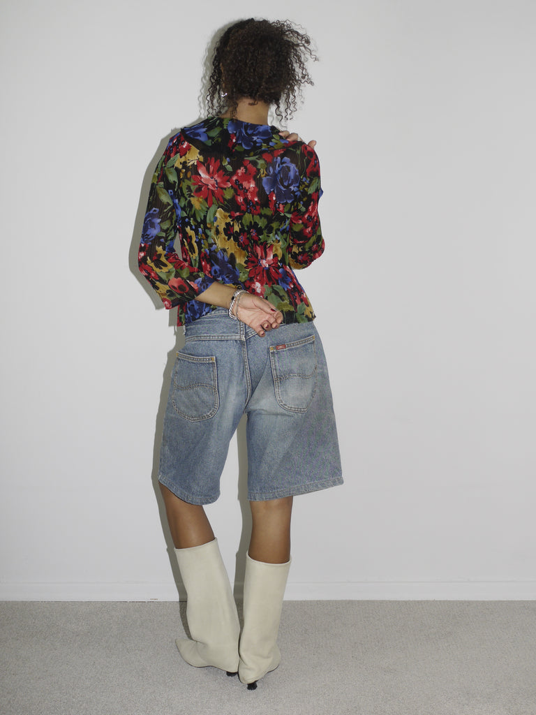 Model wearing a colorful sheer floral wrap blouse with a deep neckline, paired with loose-fitting vintage denim Bermuda shorts. Styled with cream-colored pointed cowboy boots and silver jewelry, blending romantic boho elements with early-2000s streetwear. Shot against a plain white wall