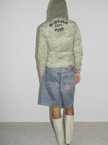 Back view of a model wearing a fitted camouflage zip-up hoodie with “G-Star 3301” branding on the upper back, paired with loose vintage denim Bermuda shorts. Styled with cream-colored pointed cowboy boots. A mix of utilitarian streetwear and Y2K western flair. Shot against a plain white background.