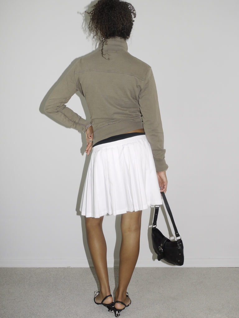 Model wearing a fitted taupe zip-up jacket with high collar and shield logo patch, paired with a white pleated mini skirt and black-and-white pointed slingback heels. Styled with a black shoulder bag and silver jewelry for a preppy-meets-sporty Y2K look. Shot against a plain white wall