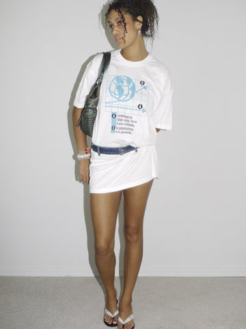 Model wearing an oversized white graphic T-shirt with blue print, styled as a mini dress and cinched at the waist with a blue belt. Paired with flip-flops, silver jewelry, and a black shoulder bag for a casual, street-style Y2K look. Shot against a plain white wall.