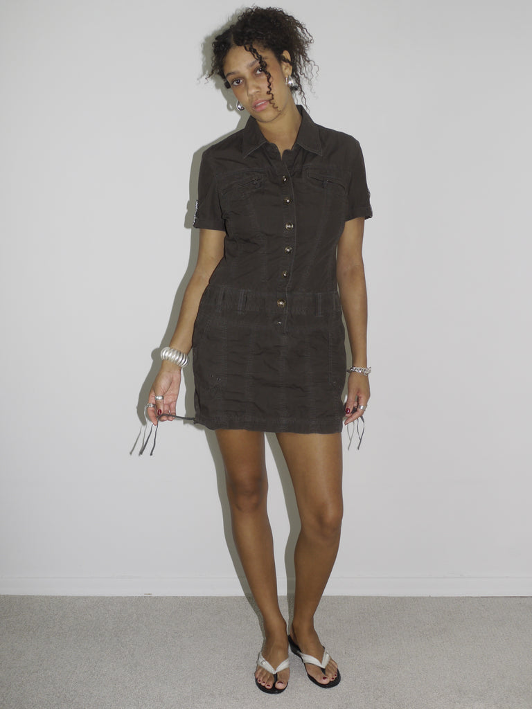Model wearing a fitted short-sleeve brown mini dress with zip and drawstring details, evoking a utilitarian, almost tactical aesthetic. Styled with flip-flops and silver accessories for a sleek, minimal look. Shot in profile against a plain white wall.