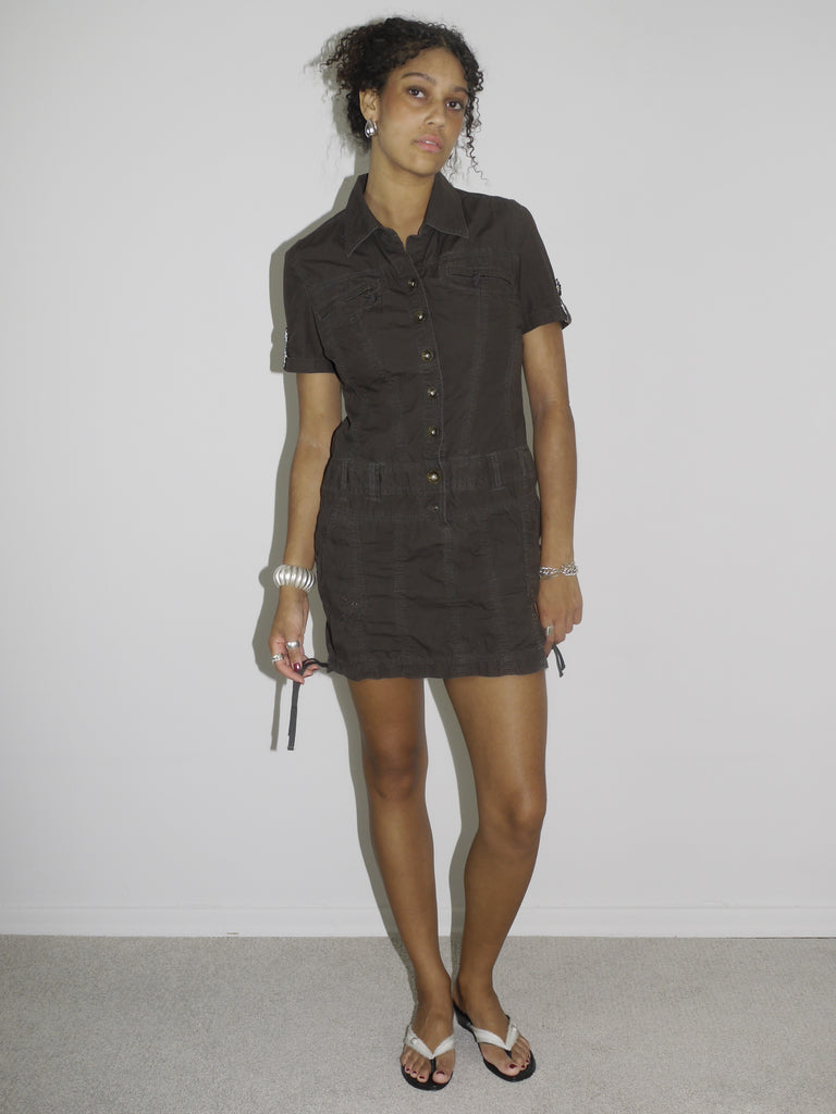 Model wearing a fitted short-sleeve brown mini dress with zip and drawstring details, evoking a utilitarian, almost tactical aesthetic. Styled with flip-flops and silver accessories for a sleek, minimal look. Shot in profile against a plain white wall.