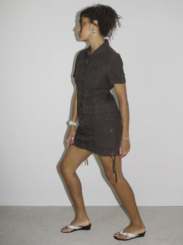 Model wearing a fitted short-sleeve brown mini dress with zip and drawstring details, evoking a utilitarian, almost tactical aesthetic. Styled with flip-flops and silver accessories for a sleek, minimal look. Shot in profile against a plain white wall.