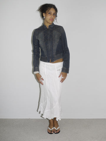 Model wearing a dark denim fitted jacket with zipper and seam detailing, paired with a flowy white crinkled midi skirt. Styled with flip-flops and subtle jewelry for a soft-meets-structured vintage look. Shot against a plain white wall.