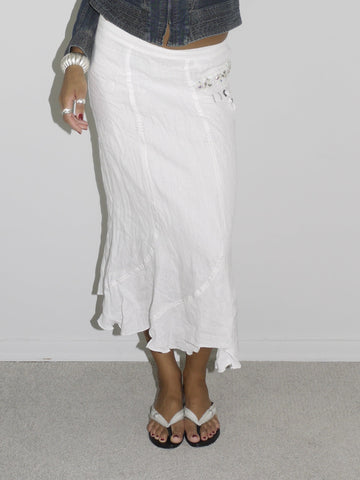 Model wearing a dark denim fitted jacket with zipper and seam detailing, paired with a flowy white crinkled midi skirt. Styled with flip-flops and subtle jewelry for a soft-meets-structured vintage look. Shot against a plain white wall.