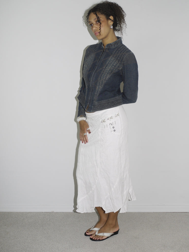 Model wearing a dark denim fitted jacket with zipper and seam detailing, paired with a flowy white crinkled midi skirt. Styled with flip-flops and subtle jewelry for a soft-meets-structured vintage look. Shot against a plain white wall.