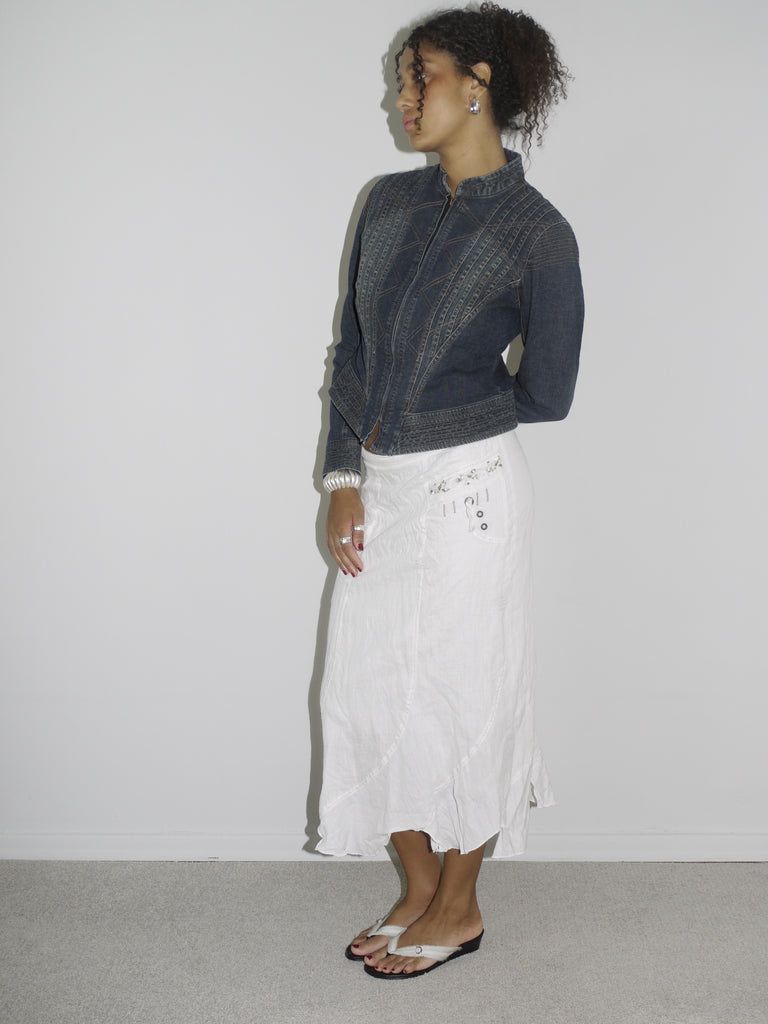 Model wearing a dark denim fitted jacket with zipper and seam detailing, paired with a flowy white crinkled midi skirt. Styled with flip-flops and subtle jewelry for a soft-meets-structured vintage look. Shot against a plain white wall.