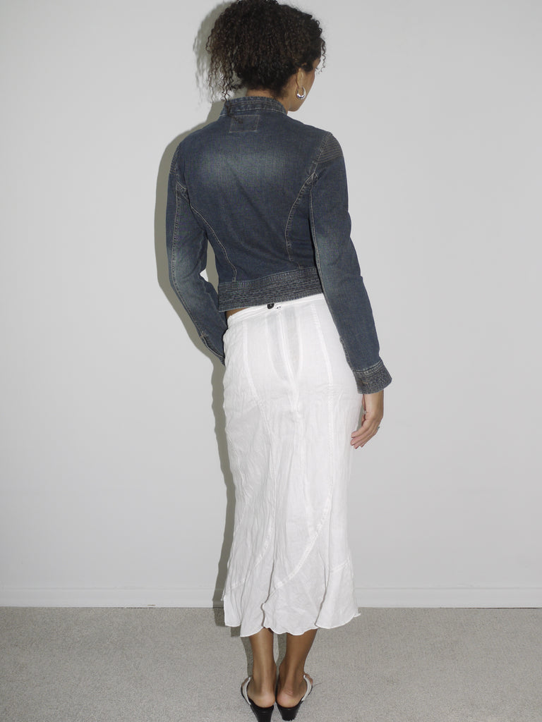 Model wearing a dark denim fitted jacket with zipper and seam detailing, paired with a flowy white crinkled midi skirt. Styled with flip-flops and subtle jewelry for a soft-meets-structured vintage look. Shot against a plain white wall.