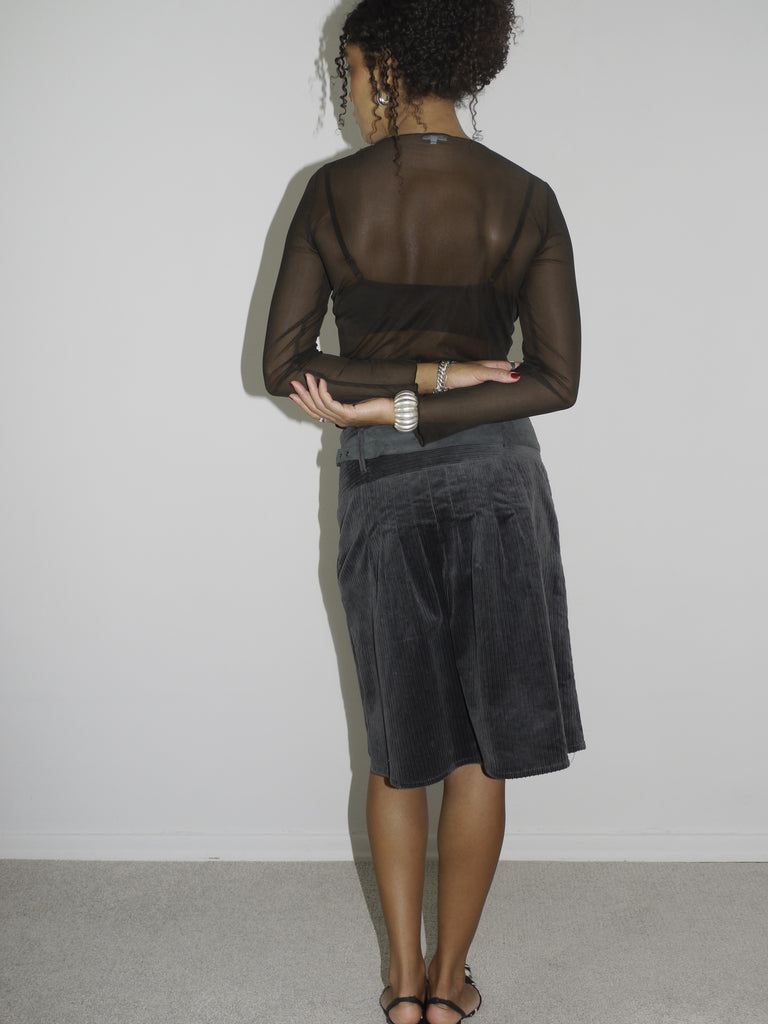 Model wearing a sheer black long-sleeve mesh top layered over a black bra, paired with a dark grey low-rise corduroy skirt featuring a wide waistband and front pocket detail. Styled with pointed black-and-white printed heels and silver jewelry for an edgy, Y2K-inspired look. Shot against a plain white wall.