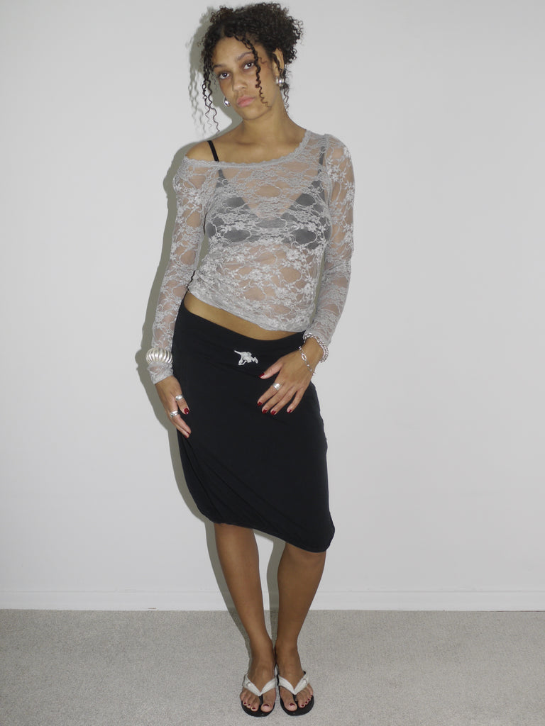 Model wearing a sheer long-sleeve lace top in light grey over a black bra, paired with a black midi skirt featuring a side brooch detail. Styled with flip-flops and silver jewelry for a feminine yet bold Y2K-inspired look. Shot against a plain white wall.