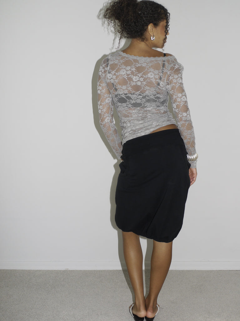 Model wearing a sheer long-sleeve lace top in light grey over a black bra, paired with a black midi skirt featuring a side brooch detail. Styled with flip-flops and silver jewelry for a feminine yet bold Y2K-inspired look. Shot against a plain white wall.