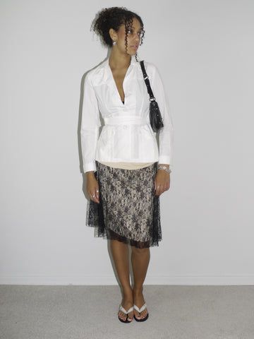 Model wearing a structured white belted blouse with a deep neckline, paired with a black lace-overlay midi skirt. Styled with flip-flops, silver jewelry, and a small black shoulder bag, creating a contrast between classic tailoring and romantic sheer textures. Shot against a plain white wall.
