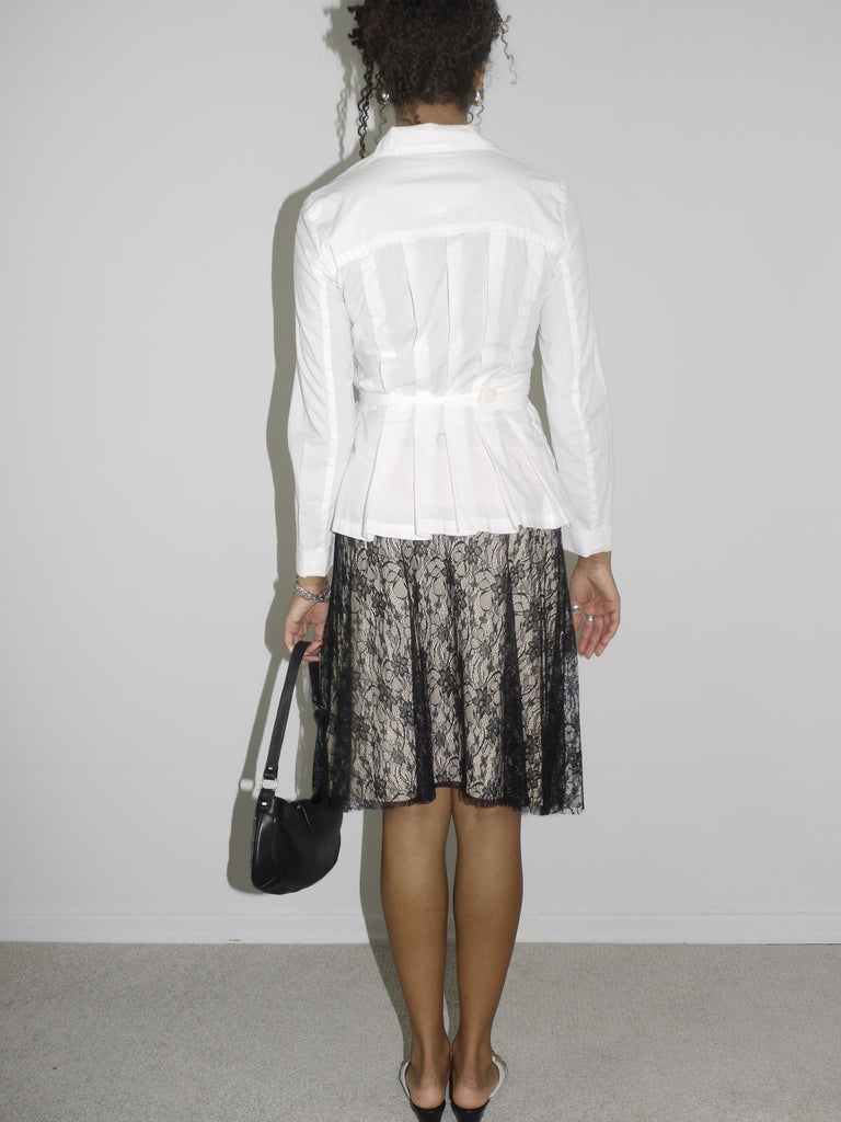 Model wearing a structured white belted blouse with a deep neckline, paired with a black lace-overlay midi skirt. Styled with flip-flops, silver jewelry, and a small black shoulder bag, creating a contrast between classic tailoring and romantic sheer textures. Shot against a plain white wall.