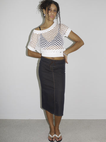 Model wearing a cropped white open-knit mesh top layered over a black bra, paired with a high-waisted dark denim midi skirt featuring contrast stitching and a wrap-effect detail. Styled with flip-flops and silver jewelry for a modern twist on early-2000s streetwear. Shot against a plain white background.