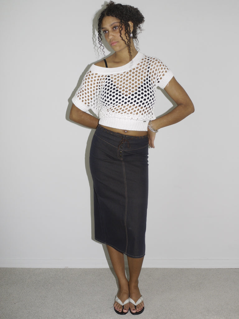 Model wearing a cropped white open-knit mesh top layered over a black bra, paired with a high-waisted dark denim midi skirt featuring contrast stitching and a wrap-effect detail. Styled with flip-flops and silver jewelry for a modern twist on early-2000s streetwear. Shot against a plain white background.